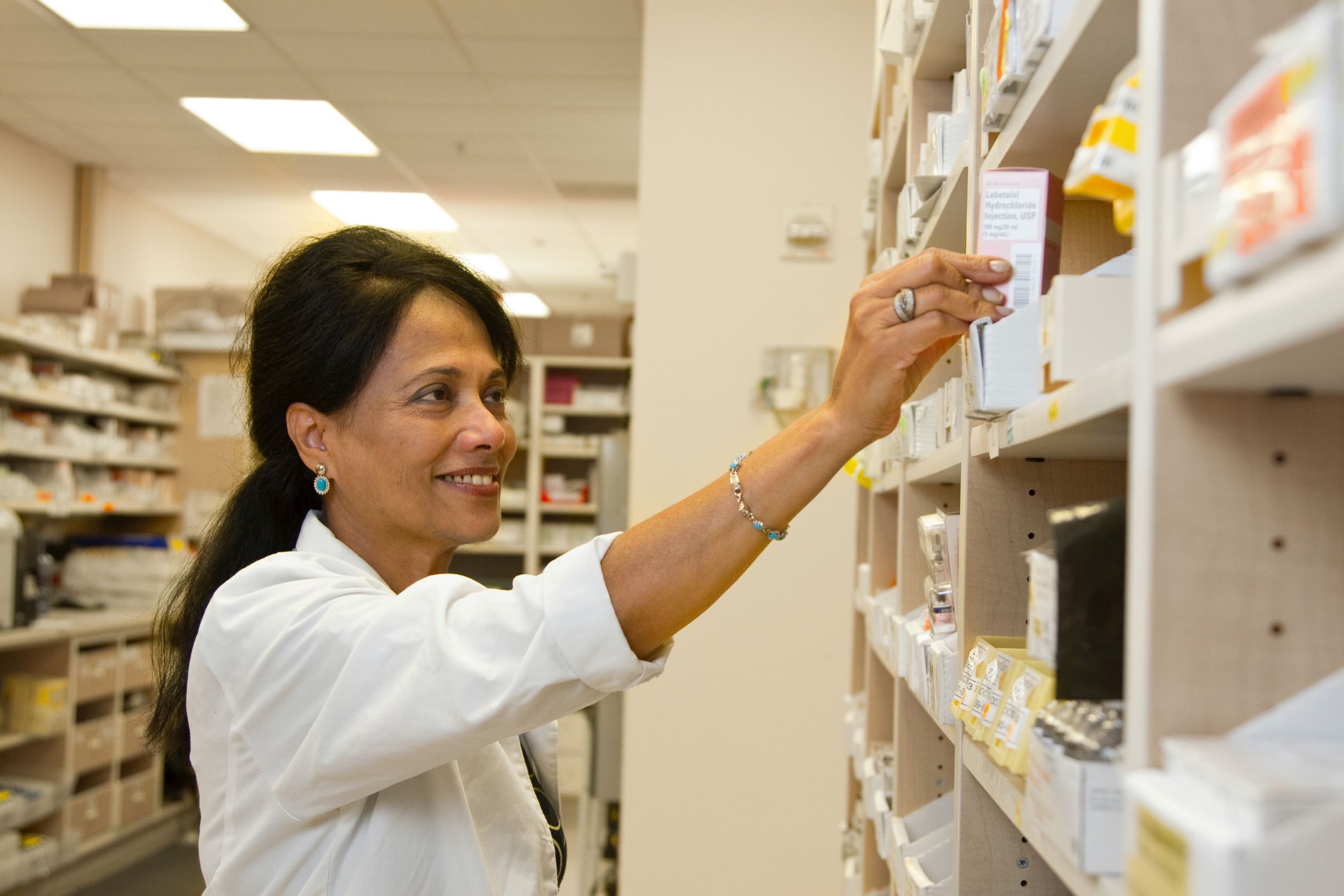 Pharmacist smiling and reaching for something on pharmacy shelf.