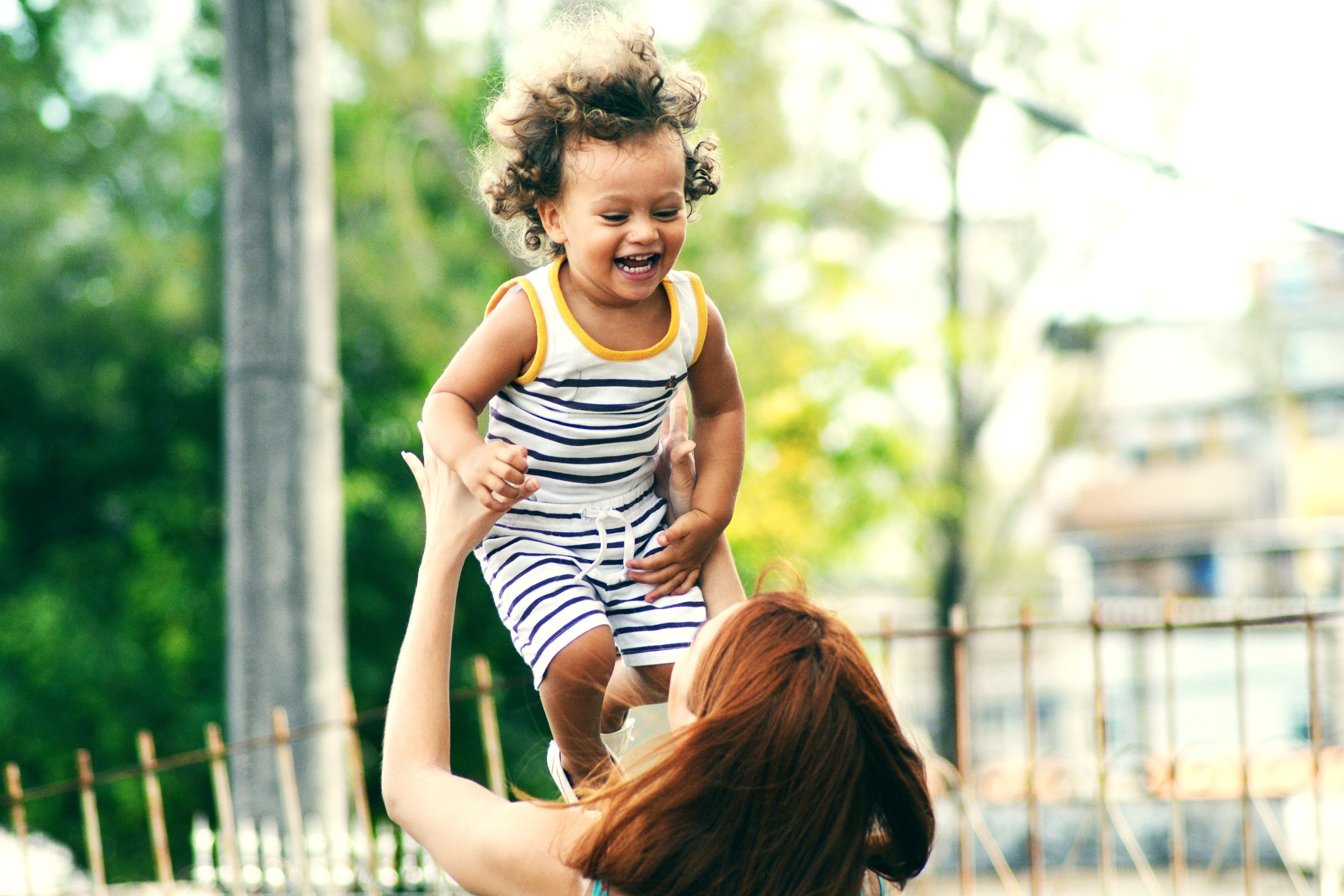 Child smiling and being held in the air by her mother outdoors.