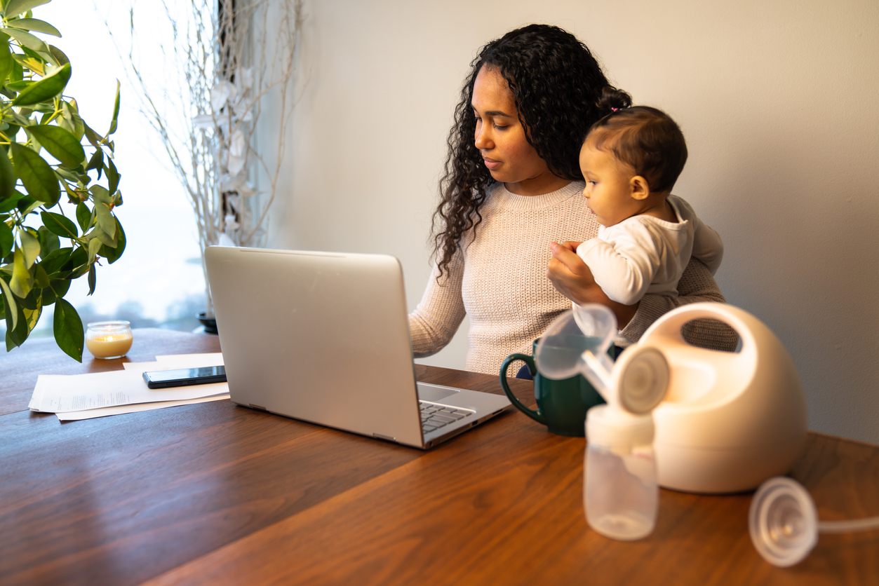 mother holds her daughter while taking notes at her dining table serving as a temporary remote work from home station with breast pump in foreground.