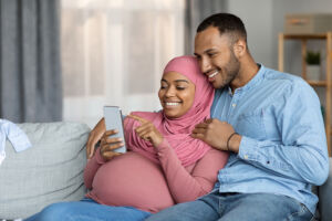 Happy Pregnant Couple Using Smartphone While Relaxing Together At Home