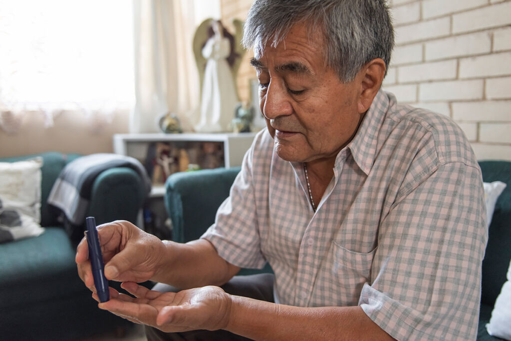 An older Hispanic man sitting on a couch at home. He is checking his blood sugar levels with a glucose monitor, focusing carefully as part of diabetes self-care and heart health management.